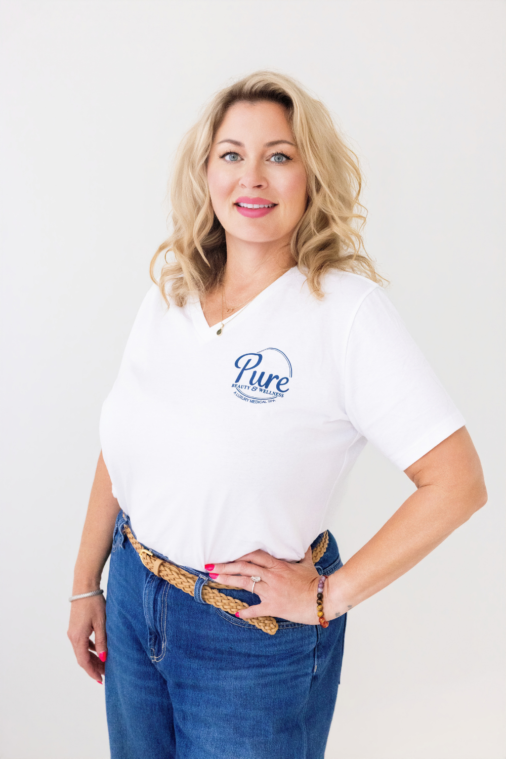 Portrait of a smiling blonde woman in a white 'pure beauty & wellness' t‑shirt and blue jeans with hands on hips.