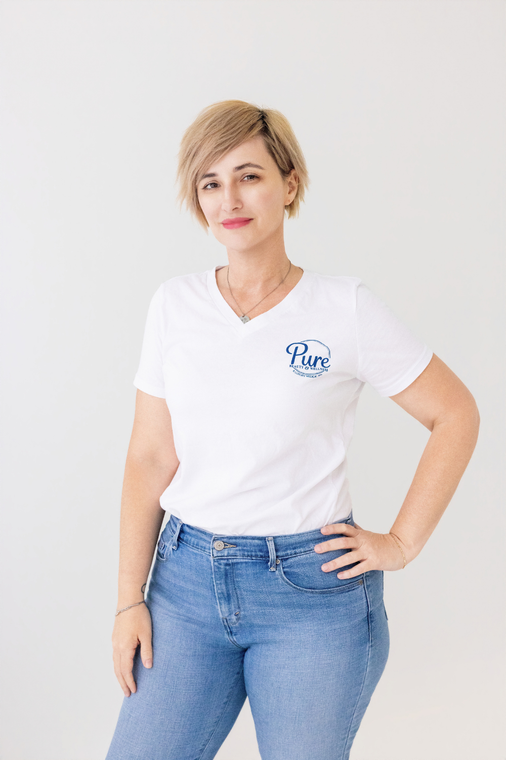 Woman with short blond hair wearing a white 'pure' logo t-shirt and blue jeans, standing with hand on hip and a slight smile.